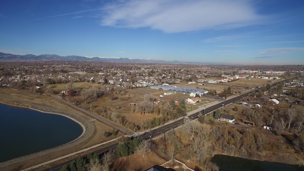 High aerial view over Westminster, Colorado with a striped road crossing beside a curved reservoir, homes, industrial buildings, distant mountains, and no visible people.