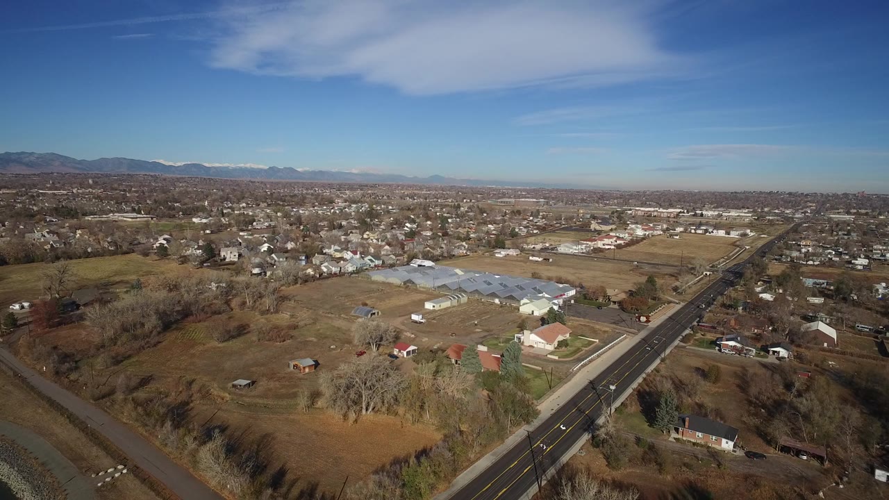 Opening aerial frame over Westminster, Colorado, with a straight road at right, scattered homes, dry fields, greenhouse roofs, distant mountains, and no visible people.