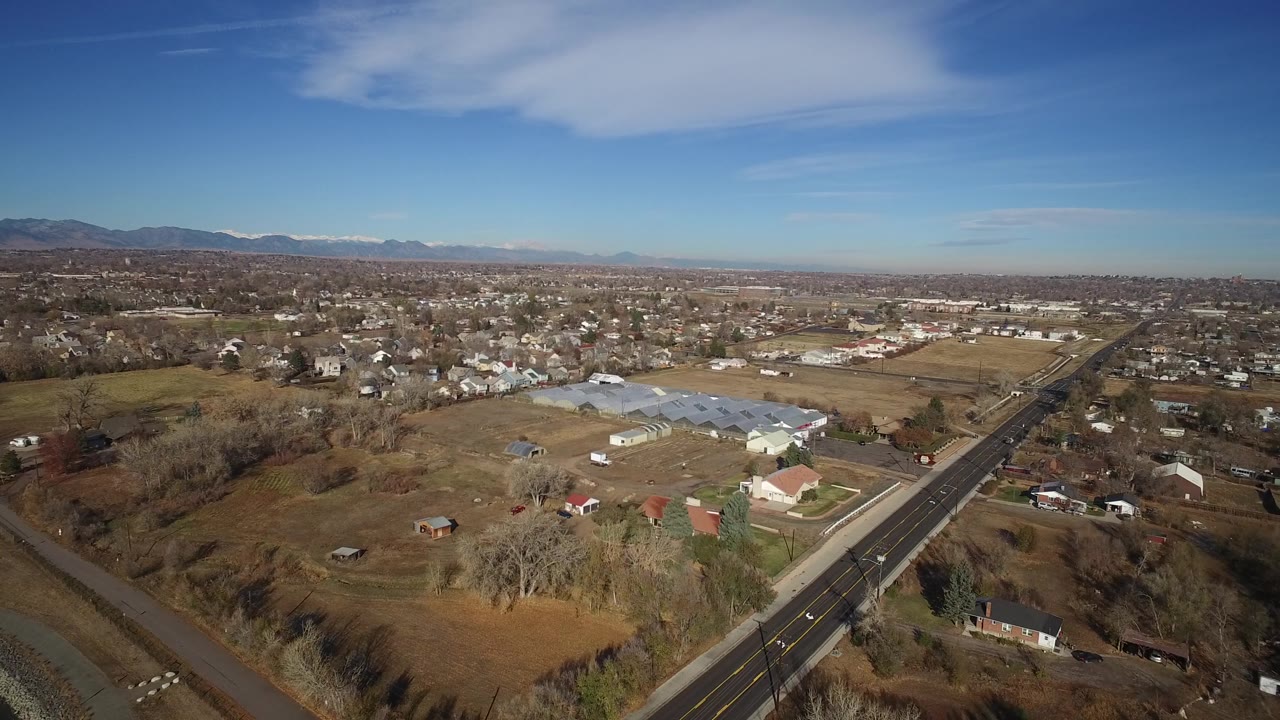 Aerial view in Westminster, Colorado, showing the road slightly right of center, houses spread across dry lots, light-colored greenhouse roofs, distant mountains, and no visible people.