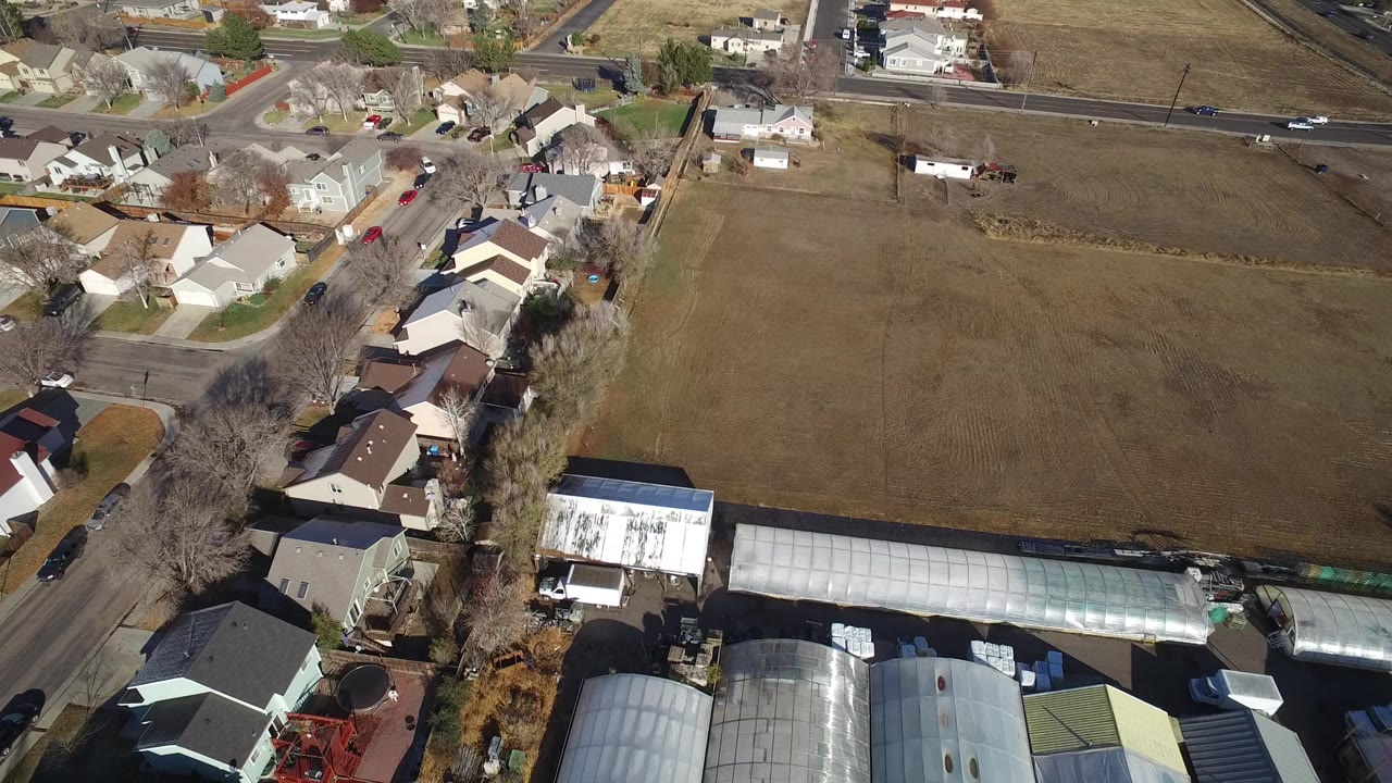 Overhead aerial view in Westminster, Colorado showing residential homes beside brown open fields, greenhouse roofs, parked cars, leafless trees, and no visible people.