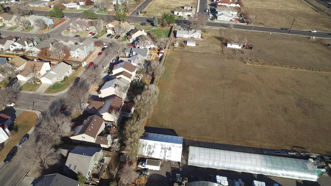 Drone-height view of homes, streets, fields, and long greenhouse buildings.