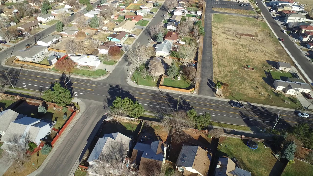 Opening aerial frame over Westminster, Colorado showing detached homes, fenced yards, a wide road, cars, and an open grass lot with no visible people.