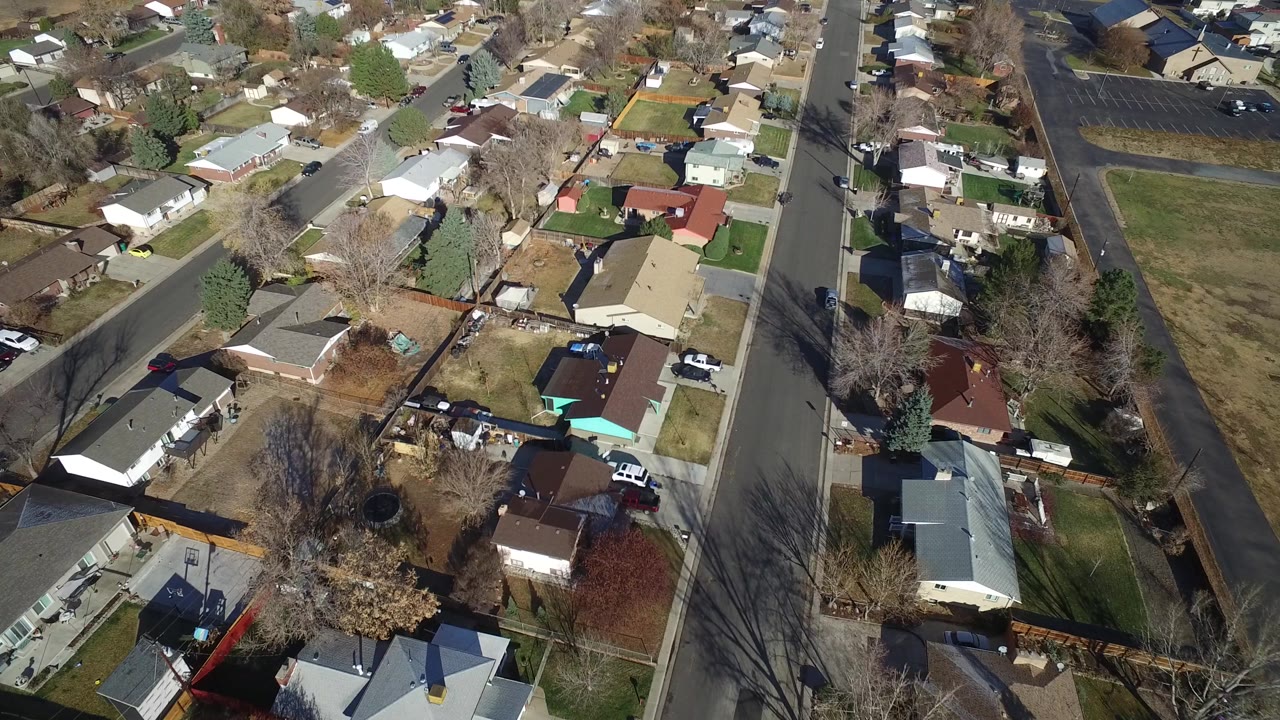 High drone view in Westminster, Colorado with a large open grassy lot on the right, clustered houses and backyards on the left, roadside trees, and no visible people.
