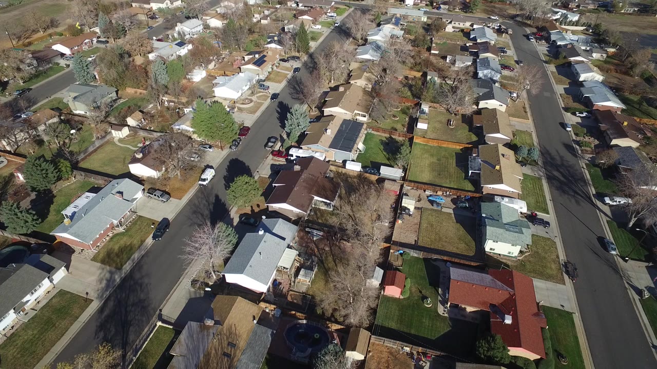 Aerial view of suburban houses, streets, yards, trees, and parked cars.