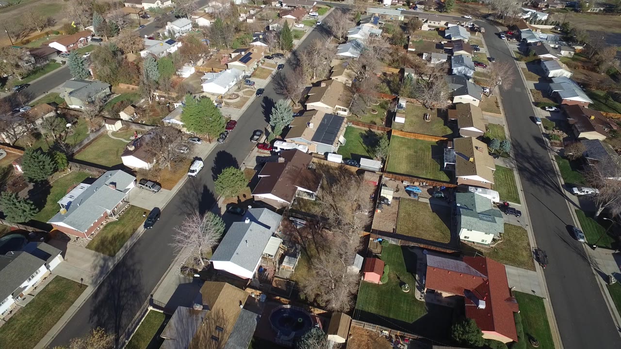 Oblique aerial frame in Westminster, Colorado showing rows of homes, a broad street on the right, winter-bare trees, green lawns, and no visible people.
