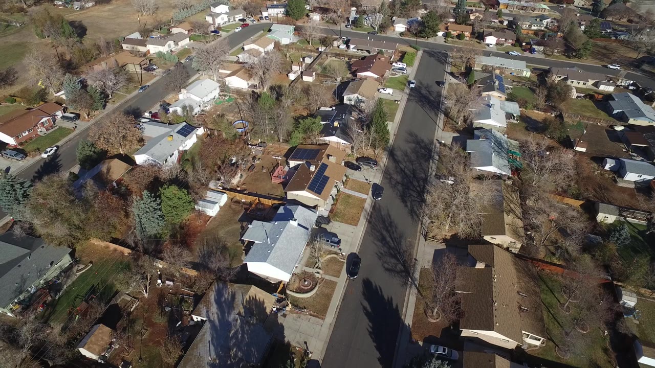 Aerial frame over Westminster, Colorado homes with a wide residential street, driveways, lawns, leafless trees, parked cars, and no visible people.