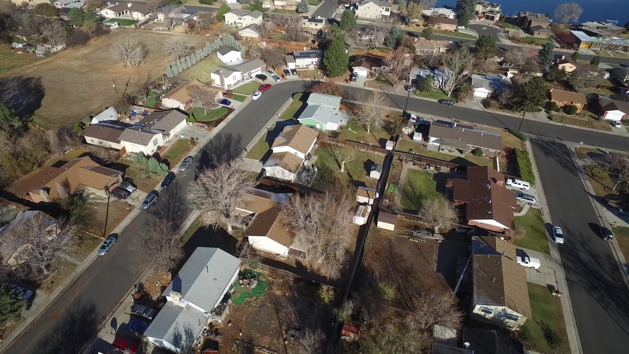 High aerial view over Westminster, Colorado homes beside a curved residential street, fenced yards, brown lawns, leafless trees, and no visible people.