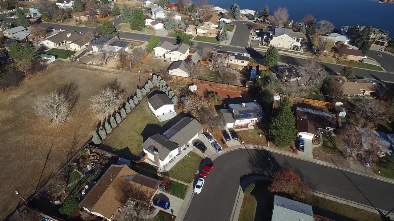 High aerial view of Westminster, Colorado houses with backyards, patios, parked cars, a residential road, and water along the top edge, with no visible people.