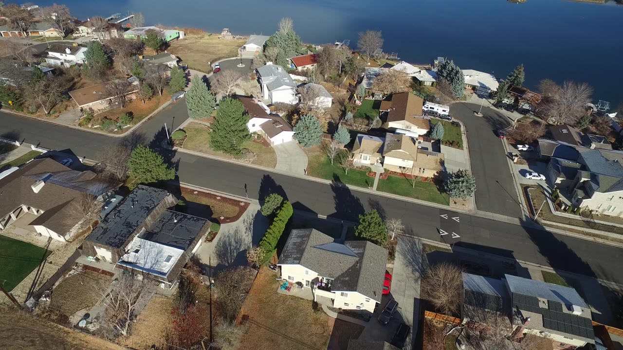 Aerial view in Westminster, Colorado with rooftops, driveways, a residential intersection, yards, trees, and the lake filling the upper background with no visible people.