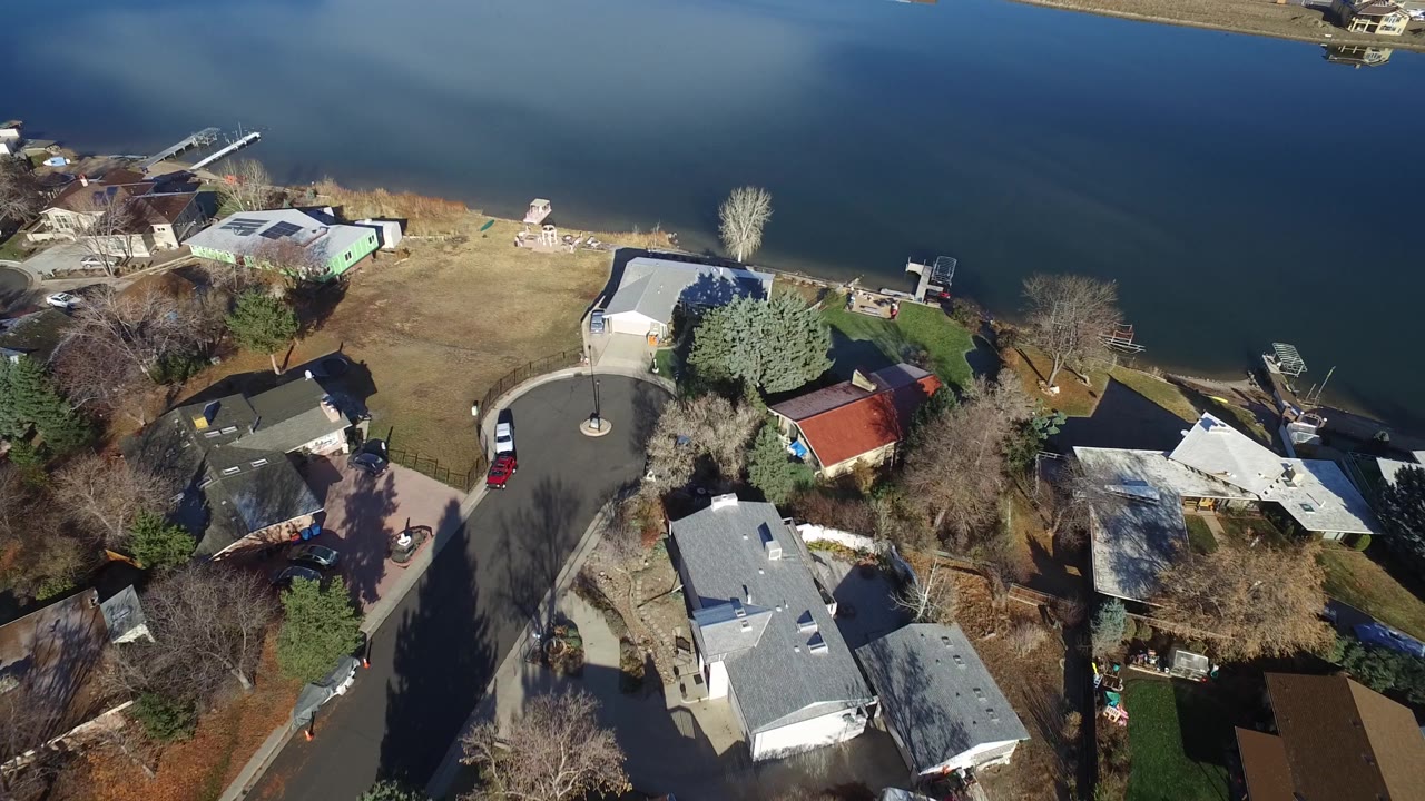 Aerial frame in Westminster, Colorado looking down on lakeside houses, a curved residential street, several parked vehicles, bare trees, small docks, and open water, with no visible people.