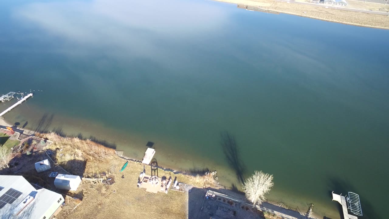 Aerial frame over a Westminster, Colorado lake with a white motorboat moving across open water, docks and private homes along the lower shoreline, and tiny people visible on the beach area.
