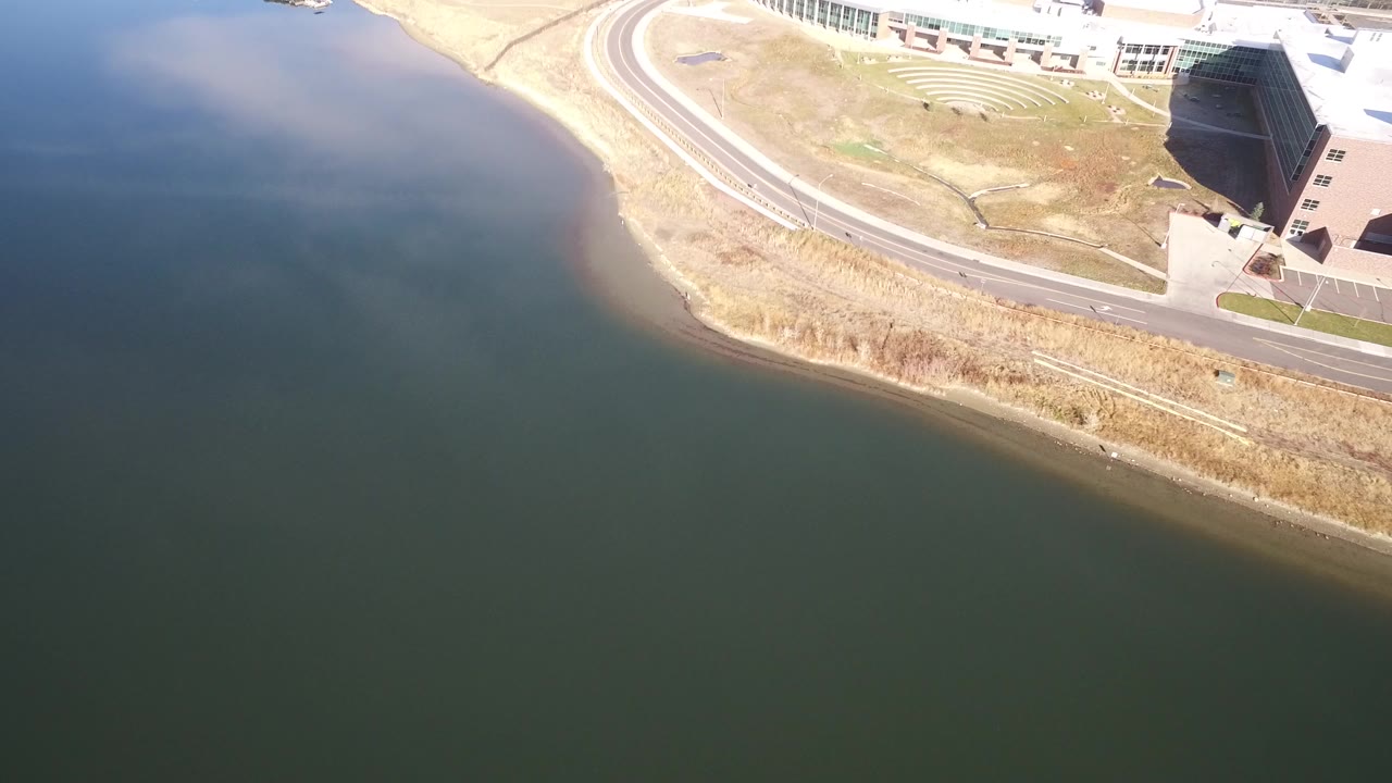 Aerial view of dark water beside a curving road and modern glass building.