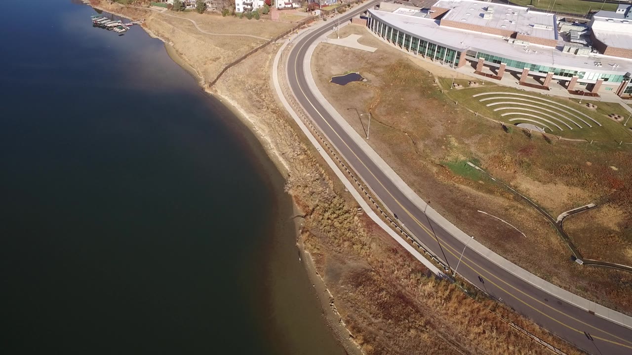 Opening overhead frame in Westminster, Colorado showing a curved empty road along dark water, tan shoreline grass, a modern glass-fronted building, and no visible people.