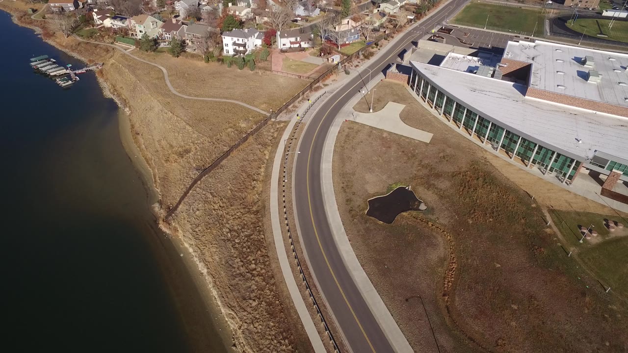 Aerial frame in Westminster, Colorado with the shoreline on the left, the curved roadway through the center, a flat-roofed modern building on the right, and homes beyond the grass.