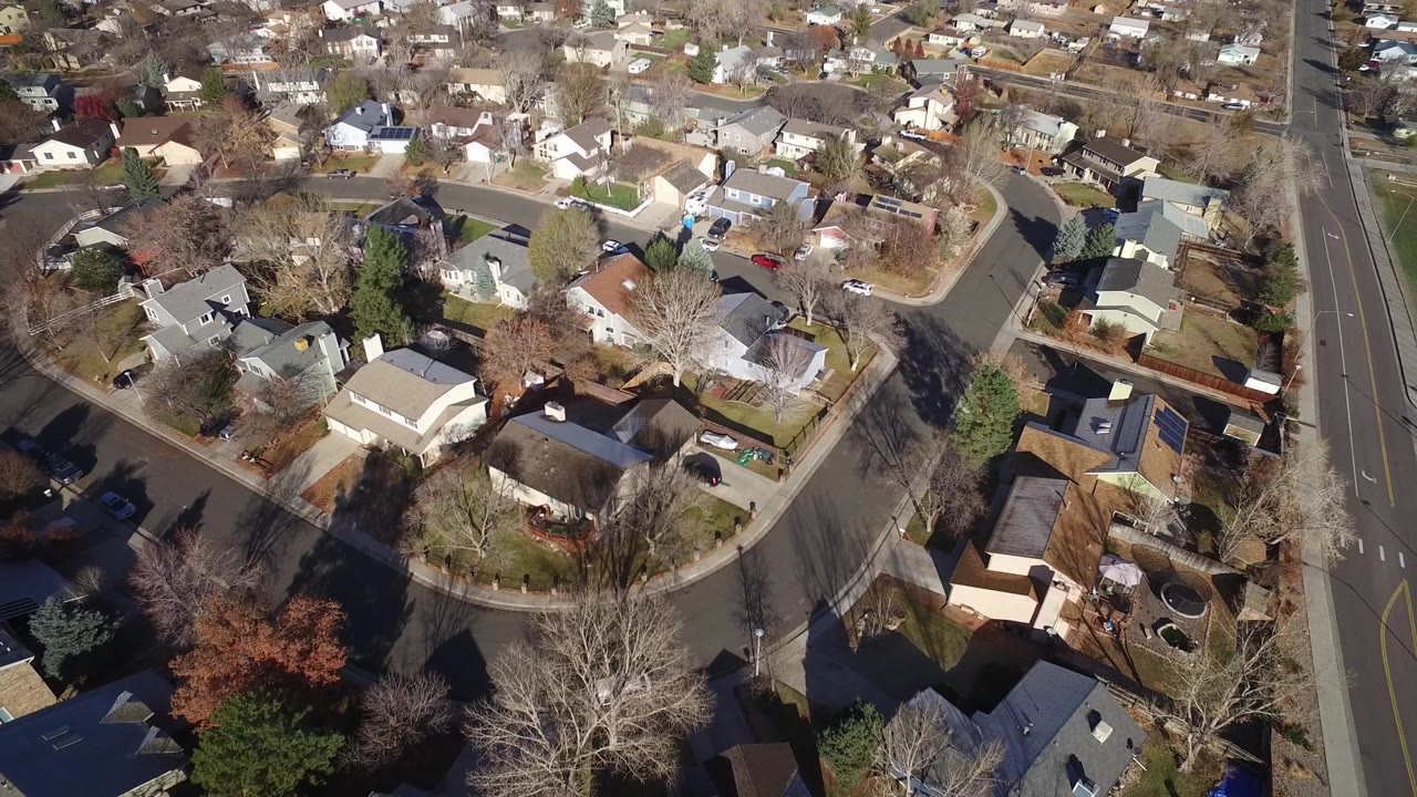 High aerial view over a Westminster, Colorado residential block with detached homes, curved streets, parked cars, bare trees, and no visible people.