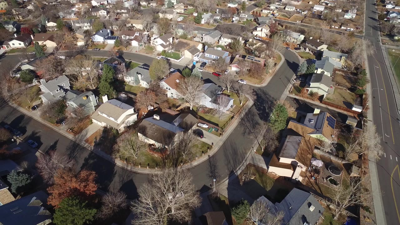 Aerial frame in Westminster, Colorado showing rooftops, a broad residential road at the right, driveways, parked vehicles, winter-bare trees, and no visible people.