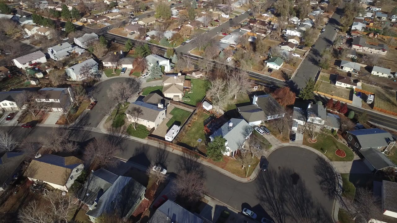 Top-down aerial frame over Westminster, Colorado homes, lawns, driveways, leafless trees, and a rounded street corner with no visible people.