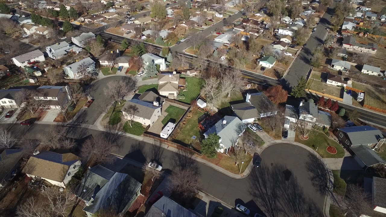 High drone view in Westminster, Colorado showing residential blocks, a broad curved road, driveways, parked vehicles, fences, and winter-bare trees with no visible people.