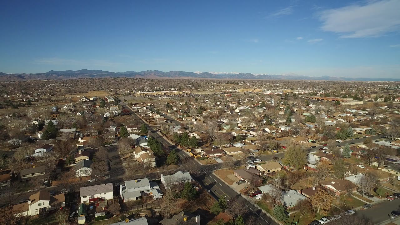Wide aerial frame over Westminster, Colorado residential streets, rooftops, leafless trees, and a distant mountain horizon under clear blue sky, with no visible people.