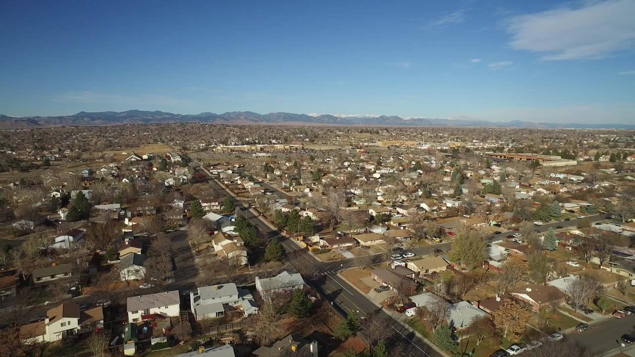 Aerial view across Westminster, Colorado homes and intersecting roads, with winter-bare trees in the foreground and low mountains across the background, with no visible people.