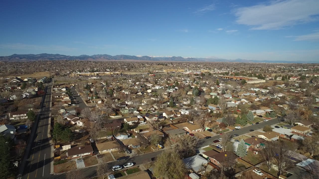 Aerial view over Westminster, Colorado, with a long straight road at left, packed houses across the frame, distant mountains, clear daylight, and no visible people.