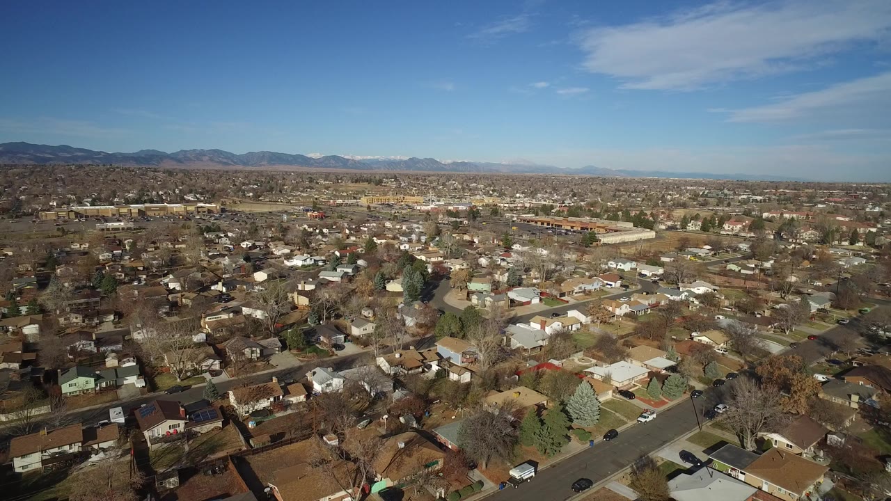 Wide aerial frame over Westminster, Colorado homes and streets with leafless trees, a blue sky, distant mountains, and no visible people.