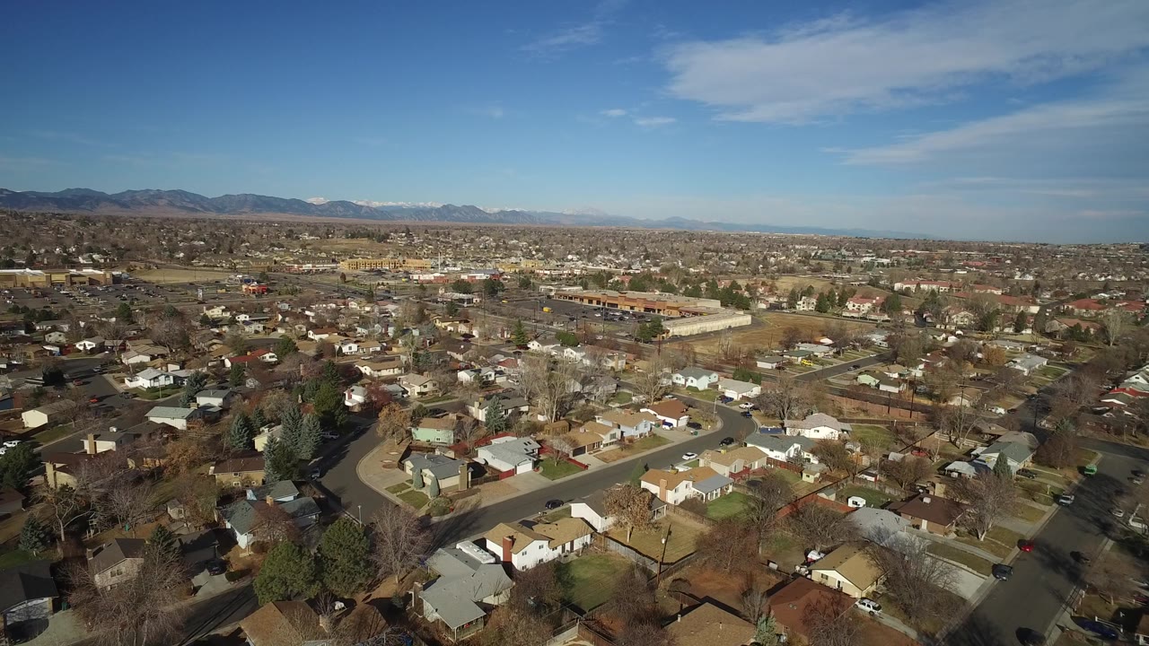 Elevated frame over Westminster, Colorado homes and curving residential roads, with bare trees, a large low building, distant mountains, and no visible people.