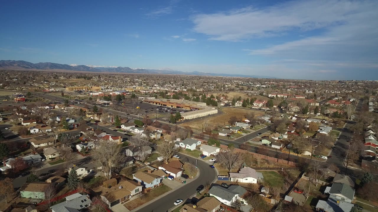 Opening aerial frame over Westminster, Colorado showing residential roofs, curved streets, bare trees, open lots, distant mountains, blue sky, and no visible people.