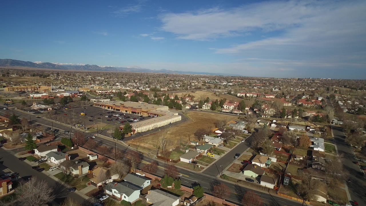 The second frame keeps a wide aerial view of Westminster, Colorado with houses in the foreground, a parking lot and commercial buildings to the left, distant foothills, and no visible people.