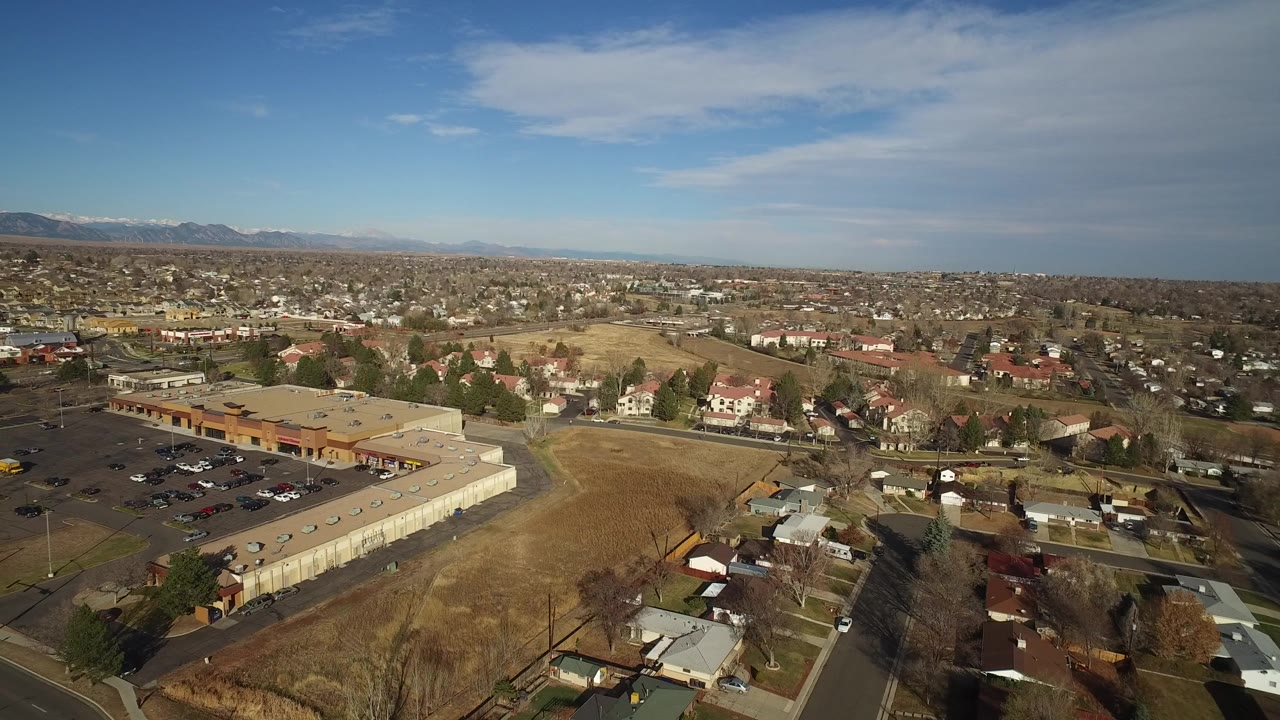 Wide aerial view over Westminster, Colorado with a tan-roofed commercial building at left, dry yellow-brown ground, suburban homes, distant mountains, blue sky, and no visible people.