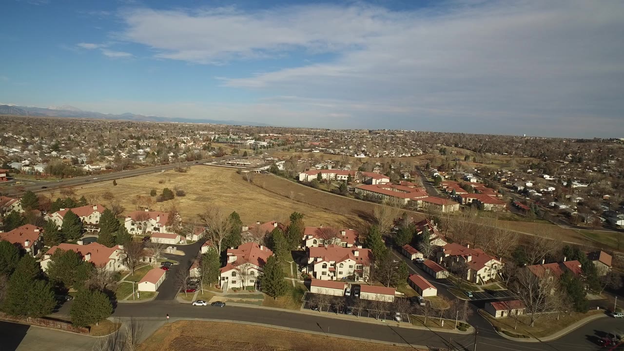 Aerial view over red-roofed homes, open field, road, suburbs, mountains, and blue sky.
