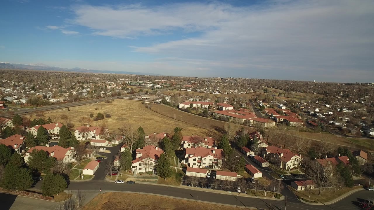 Aerial view in Westminster, Colorado with the foreground homes slightly lower in frame, the open field and road centered, and distant neighborhoods stretching toward the mountains with no visible people.