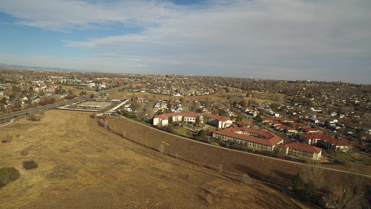 Opening aerial frame over Westminster, Colorado, with dry open fields in front, red-roofed residential buildings at lower right, roads to the left, distant houses, and no visible people.