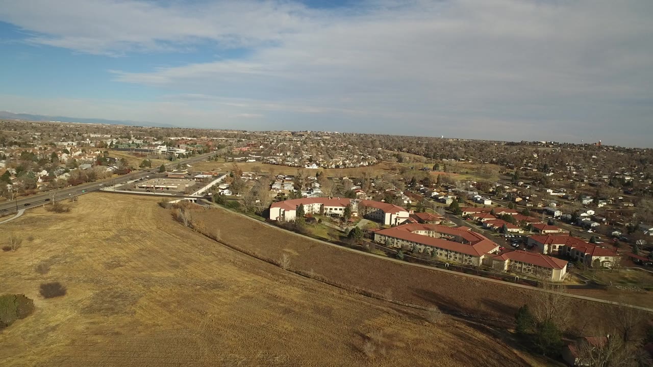 A second aerial frame in Westminster, Colorado, shows the dry field occupying more of the lower left, red-roofed buildings along the lower right, a road corridor at left, distant housing, and no visible people.
