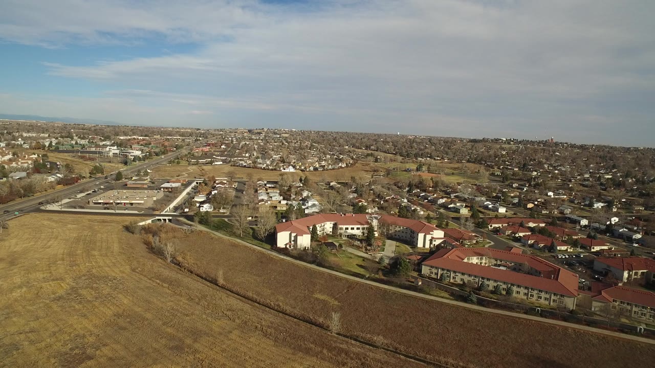 A high aerial view in Westminster, Colorado looks across dry grassland toward red-roof apartment buildings, roads, and low-rise neighborhoods, with no visible people.