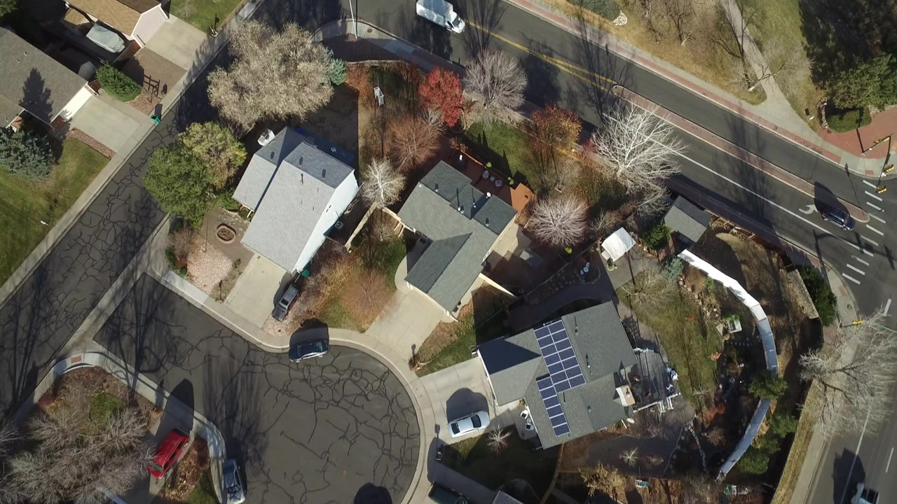 High aerial frame in Westminster, Colorado showing rooftops, a driveway, bare and flowering trees, a two-lane road, and parked vehicles, with no visible people.