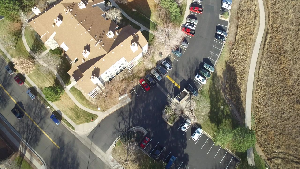 Overhead drone frame in Westminster, Colorado showing tan residential rooftops, a curved street lined with parked cars, sidewalks, bare trees, and no visible people.