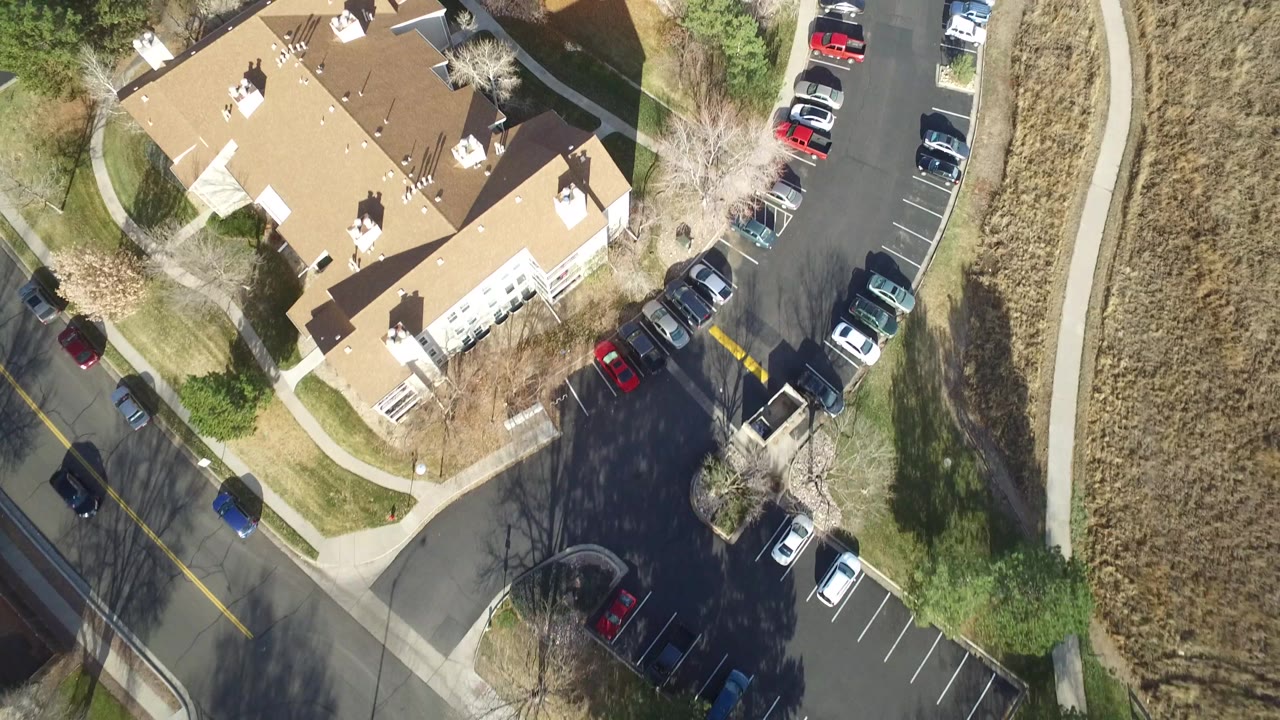 Westminster, Colorado aerial frame with the residential roofs centered, cars parked along the right side of the street, a sidewalk beside dry grass, and no visible people.
