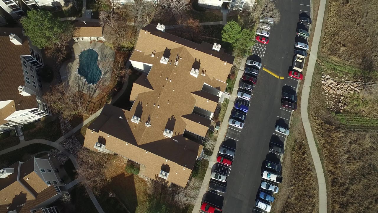 Overhead view of apartment roofs, parked cars, trees, and a small pond.