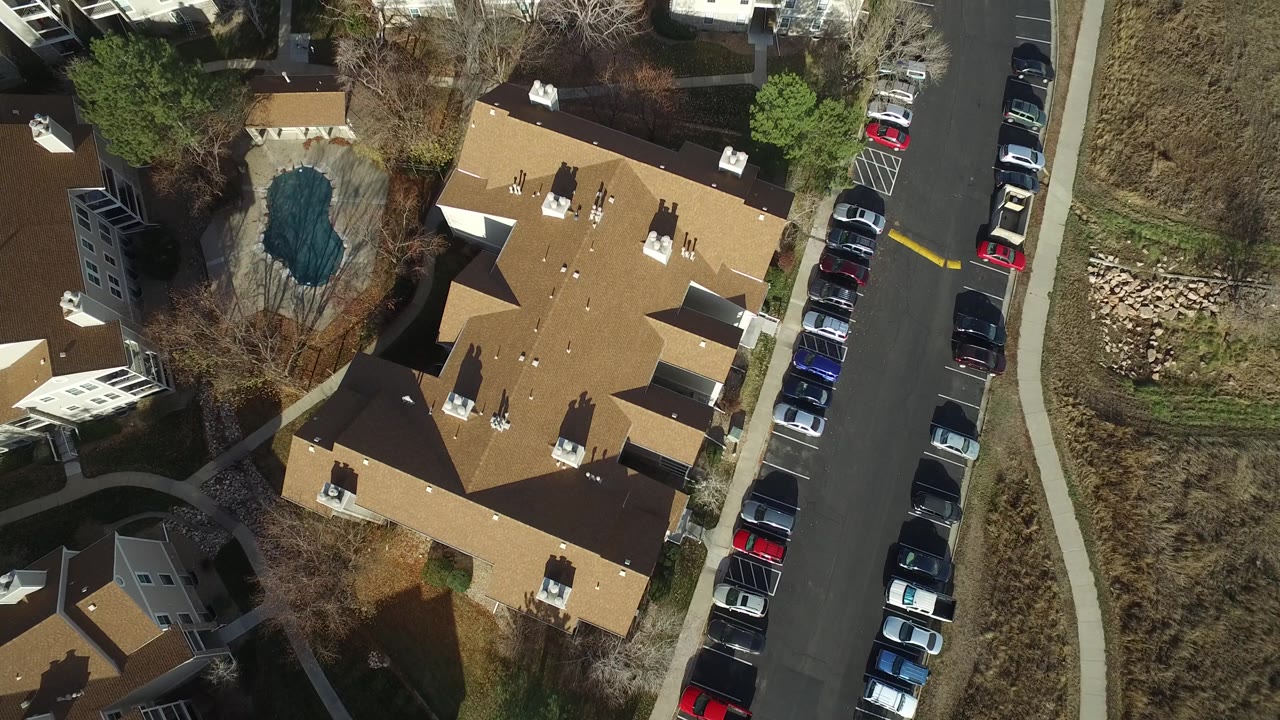 Aerial view in Westminster, Colorado showing apartment rooftops, a line of parked cars, a curving sidewalk, bare trees, open grassland, and no recognizable people visible.