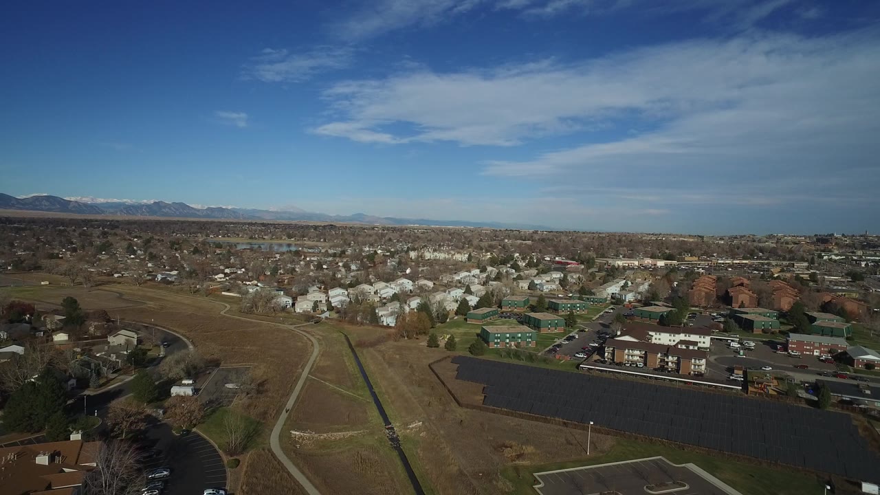 This Westminster, Colorado aerial frame keeps the dark panel rows in the lower foreground, with light-colored houses, low green-roofed buildings, distant mountains, and no visible people.