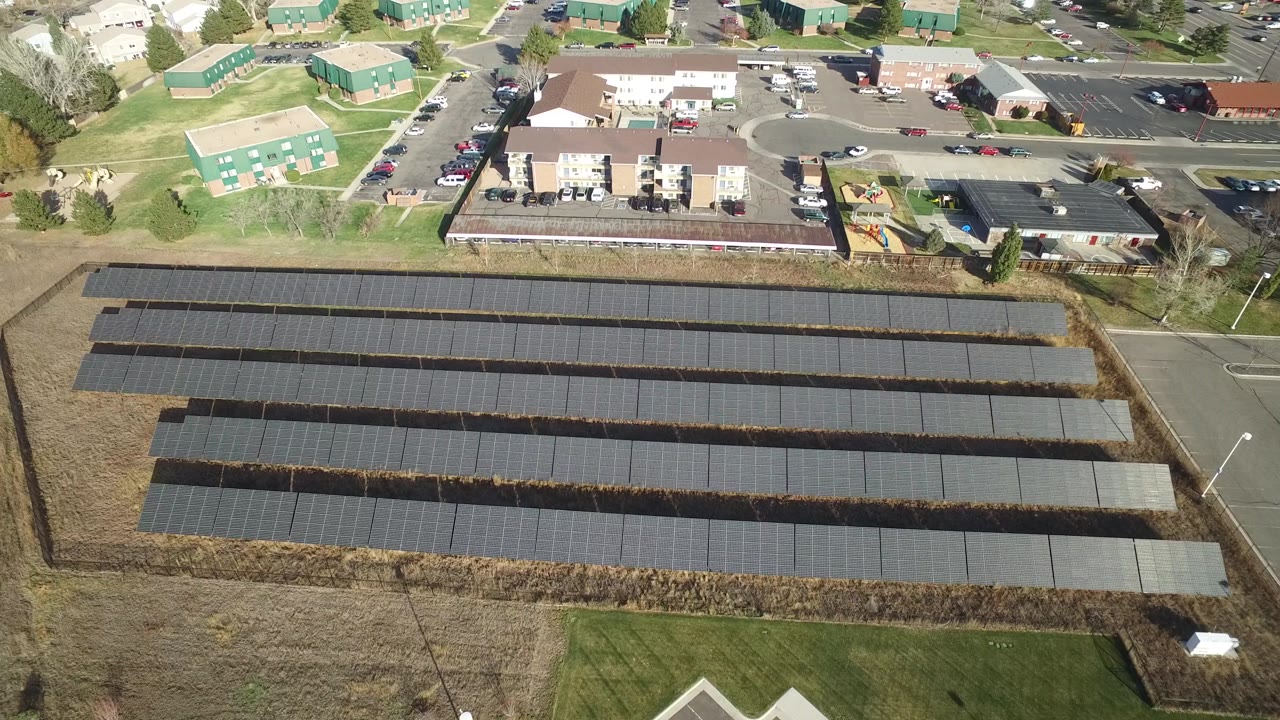 Elevated Westminster, Colorado view with the solar panel rows centered below apartment buildings, parking lots, roads, lawns, and no visible people.