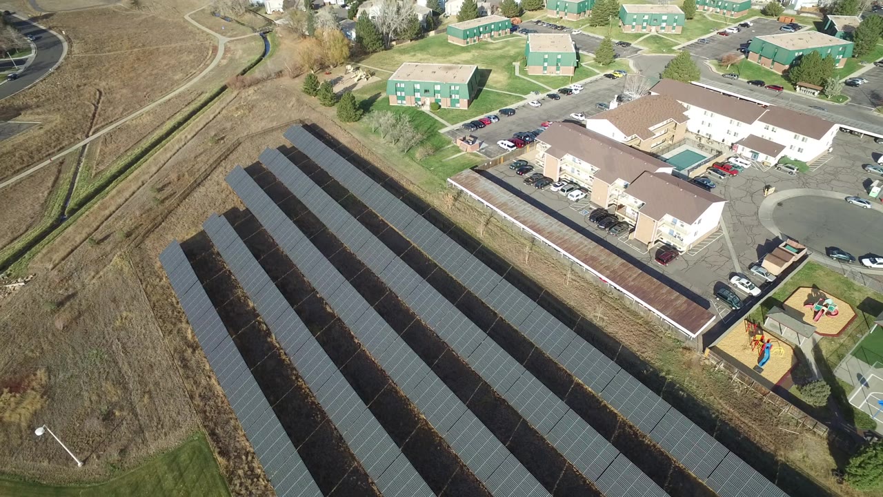 Aerial frame in Westminster, Colorado, with the solar panel rows centered lower in view and green apartment buildings, roads, cars, and open brown grassland beyond, with no visible people.