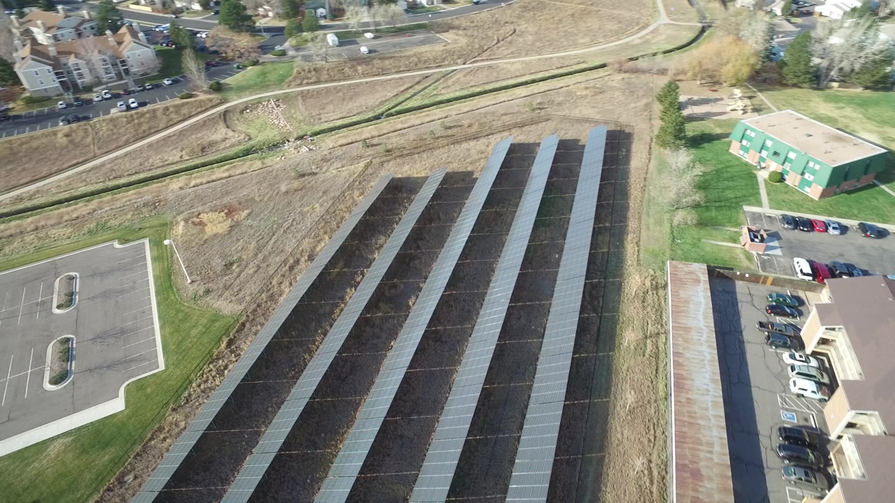 Aerial frame in Westminster, Colorado with the dark ground-mounted rows centered, parked cars along the building edge, tan open ground, and no visible people.