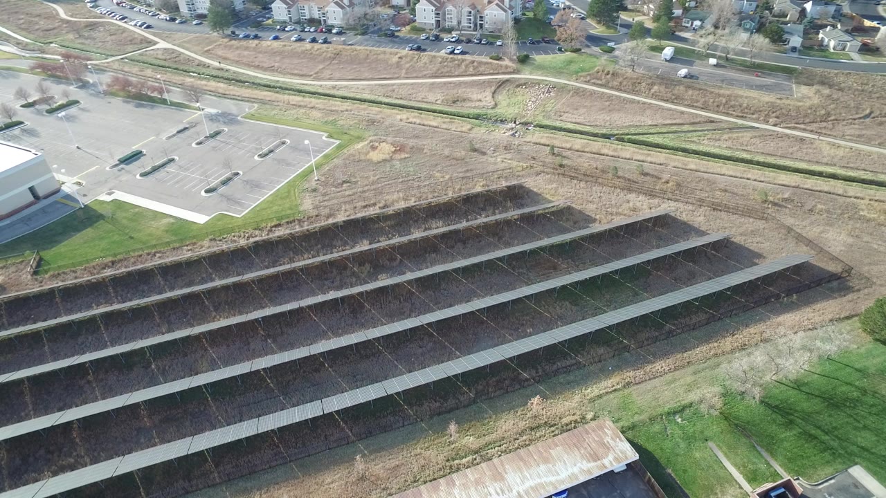 Opening frame of a Westminster, Colorado aerial view showing an empty parking lot at upper left, dark terraced drainage slopes across the center, road and buildings beyond, and no visible people.