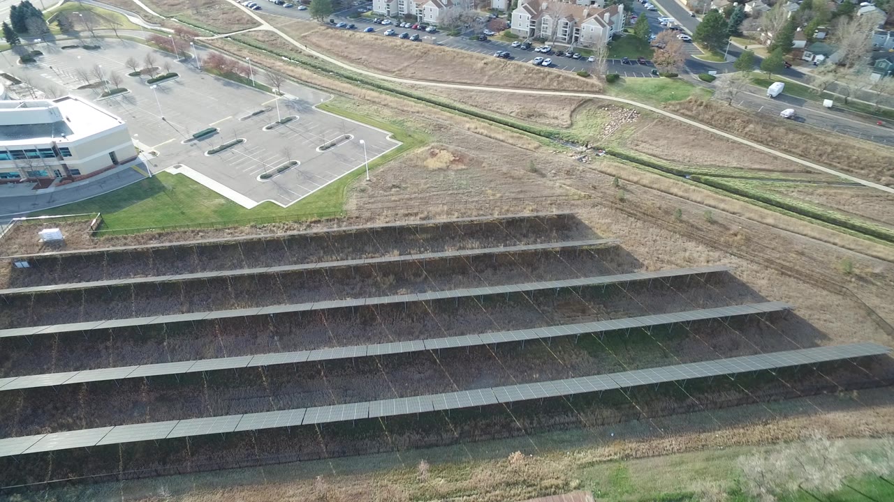 Aerial frame in Westminster, Colorado looking across dark solar panel rows toward an empty parking lot, nearby apartments, curving roads, and no visible people.