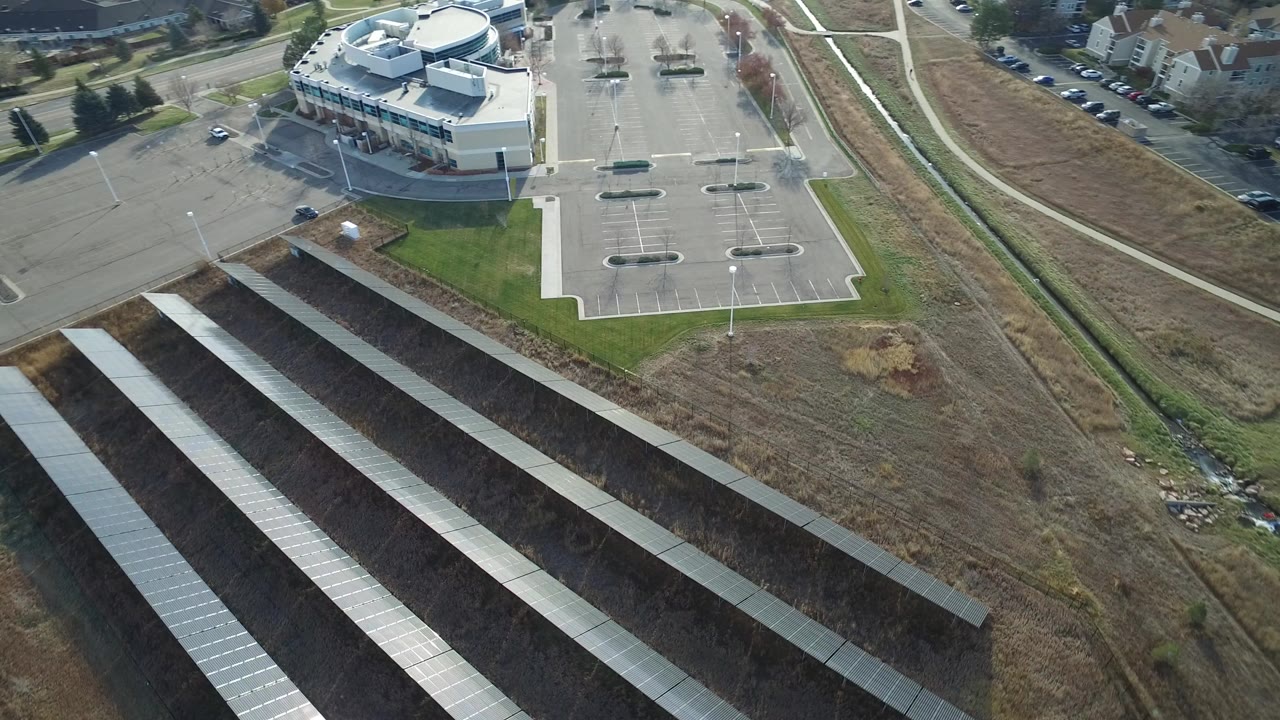 Oblique aerial view in Westminster, Colorado, with long solar panel rows in the foreground, a commercial building and parking lot above them, roads and homes beyond, and no visible people.