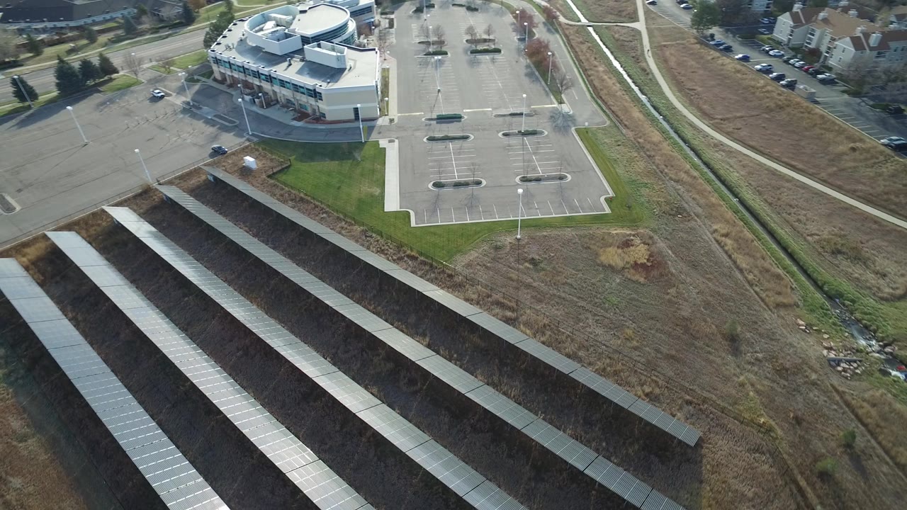 Aerial frame over Westminster, Colorado, showing multiple parallel solar panel rows across brown ground, with a parking lot, commercial building, roadways, and no visible people.