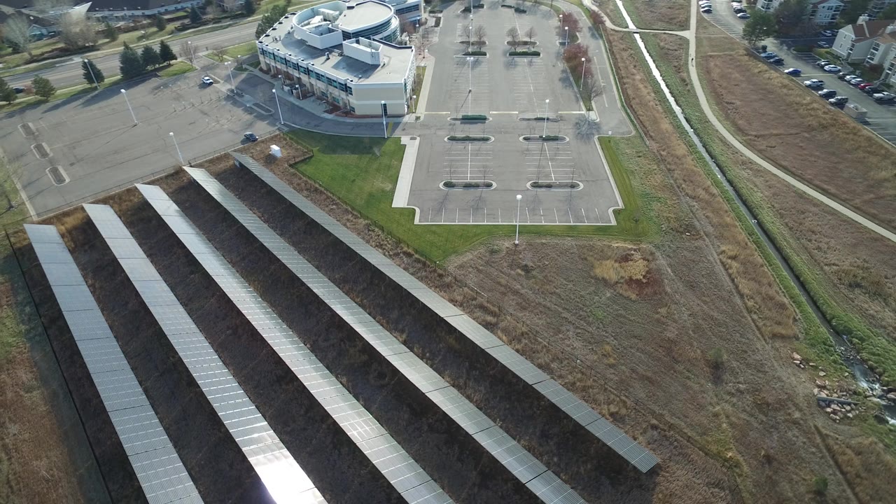 Reflective solar panels line a field below a commercial building and roads.