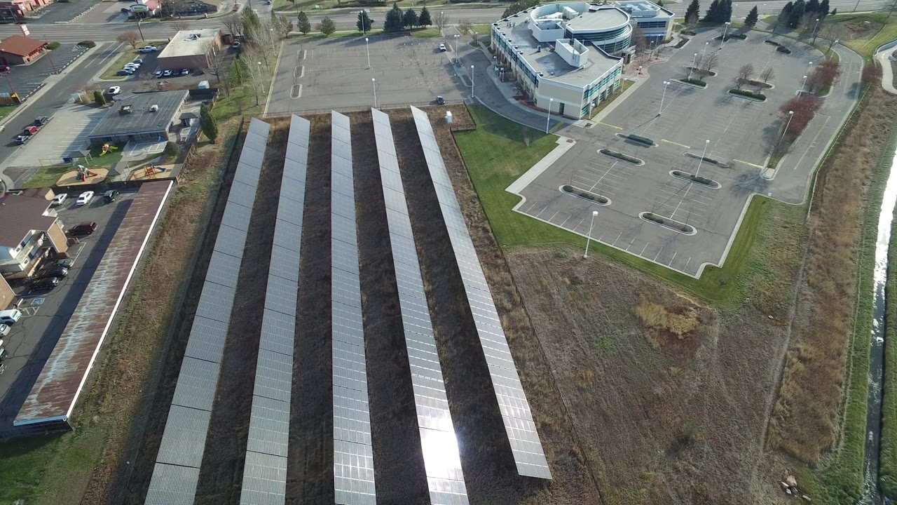 High aerial view in Westminster, Colorado showing long ground-mounted solar panel rows beside a mostly empty parking lot, low-rise buildings, roads, grass, and no visible people.